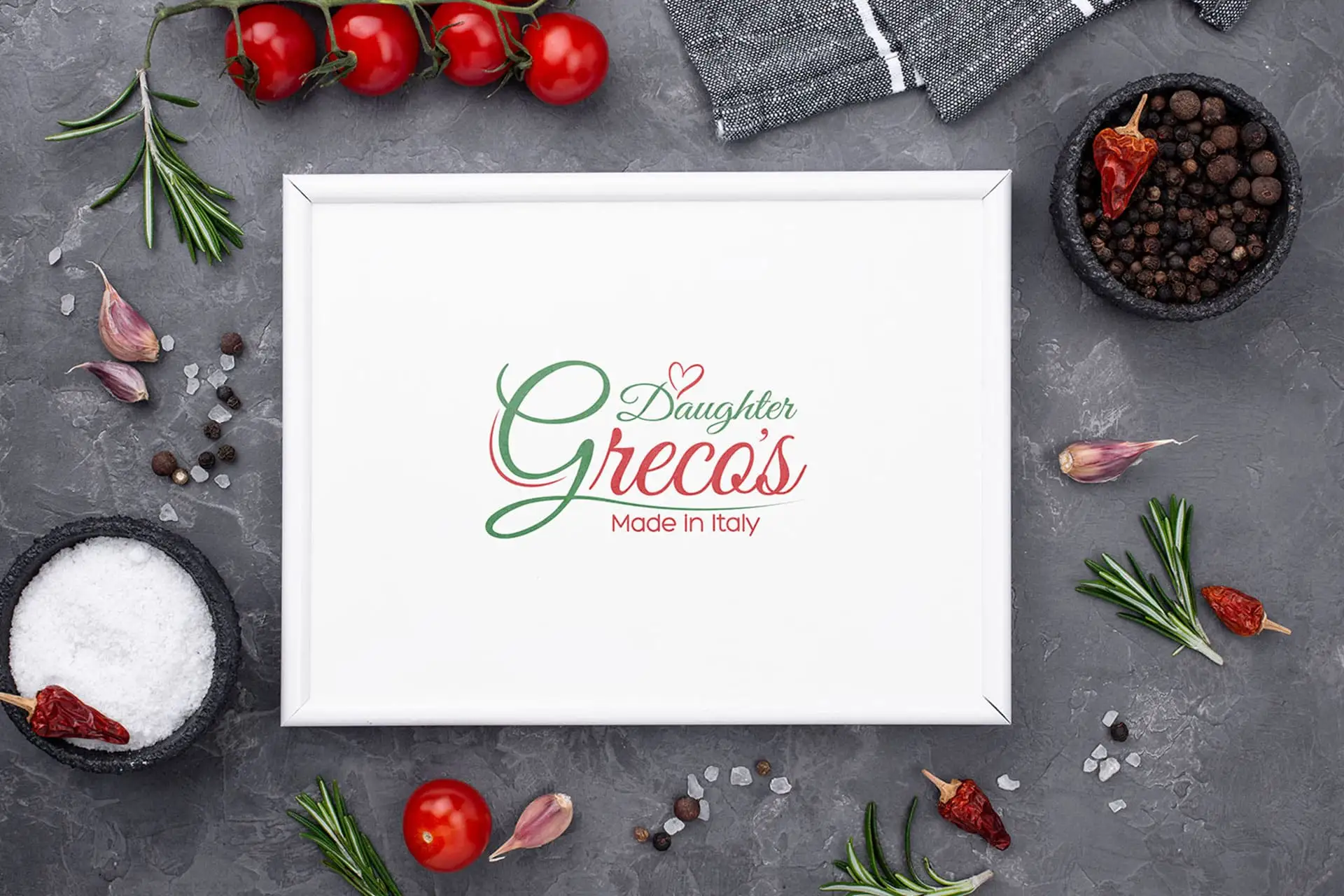 Daughter Greco's Italian Restaurant Logo Mockup: The logo, styled in elegant red and green script, is displayed on a white frame, surrounded by fresh Italian ingredients including cherry tomatoes on the vine, rosemary sprigs, garlic, and coarse salt. The tagline reads "Made in Italy," emphasizing authentic Italian cuisine and restaurant branding.