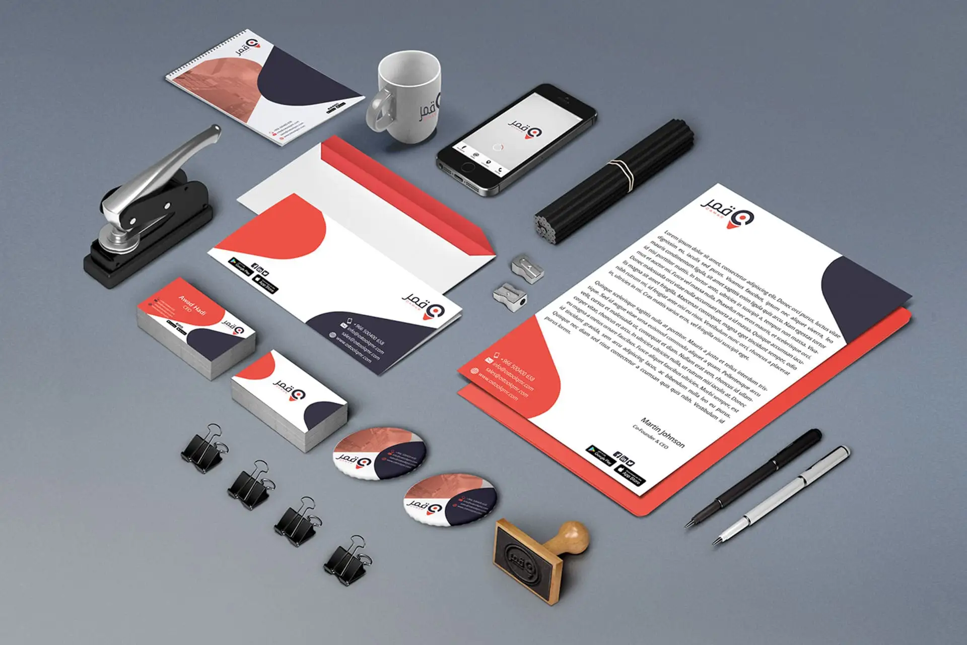 Qamar Corporate Identity Stationery Mockup: A well-organized flat lay of professional office and branding items, including letterheads, business cards, envelopes, a notebook, and a mug, all featuring the Qamar logo and a modern red, dark gray, and white geometric design. Corporate branding and professional visual identity set.