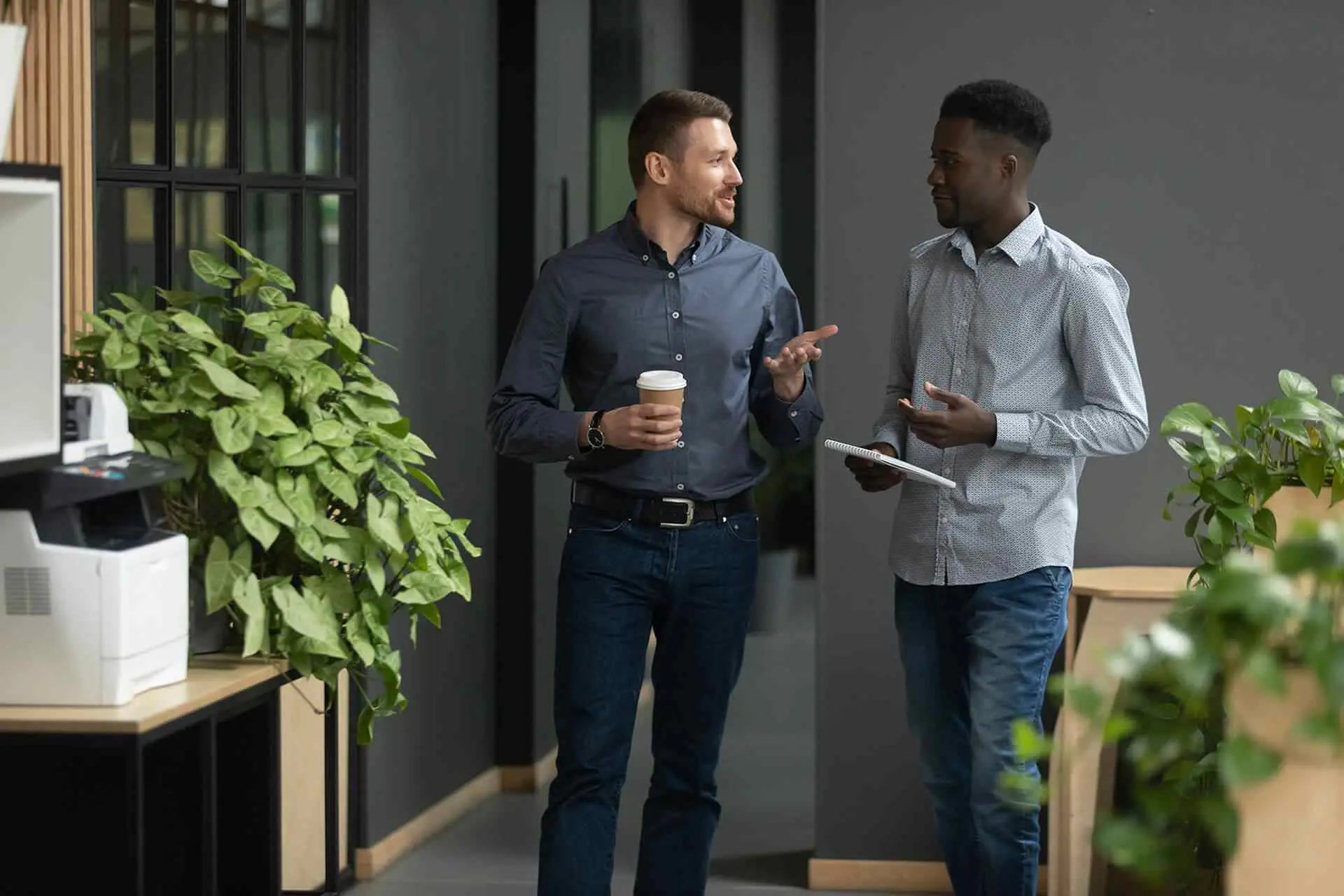 Two businessmen walking and talking in an office setting. One man is holding a coffee cup, and the other is holding a notepad, suggesting a casual but professional discussion, teamwork, or mentorship in a modern corporate hallway. The background includes interior plants and office equipment.